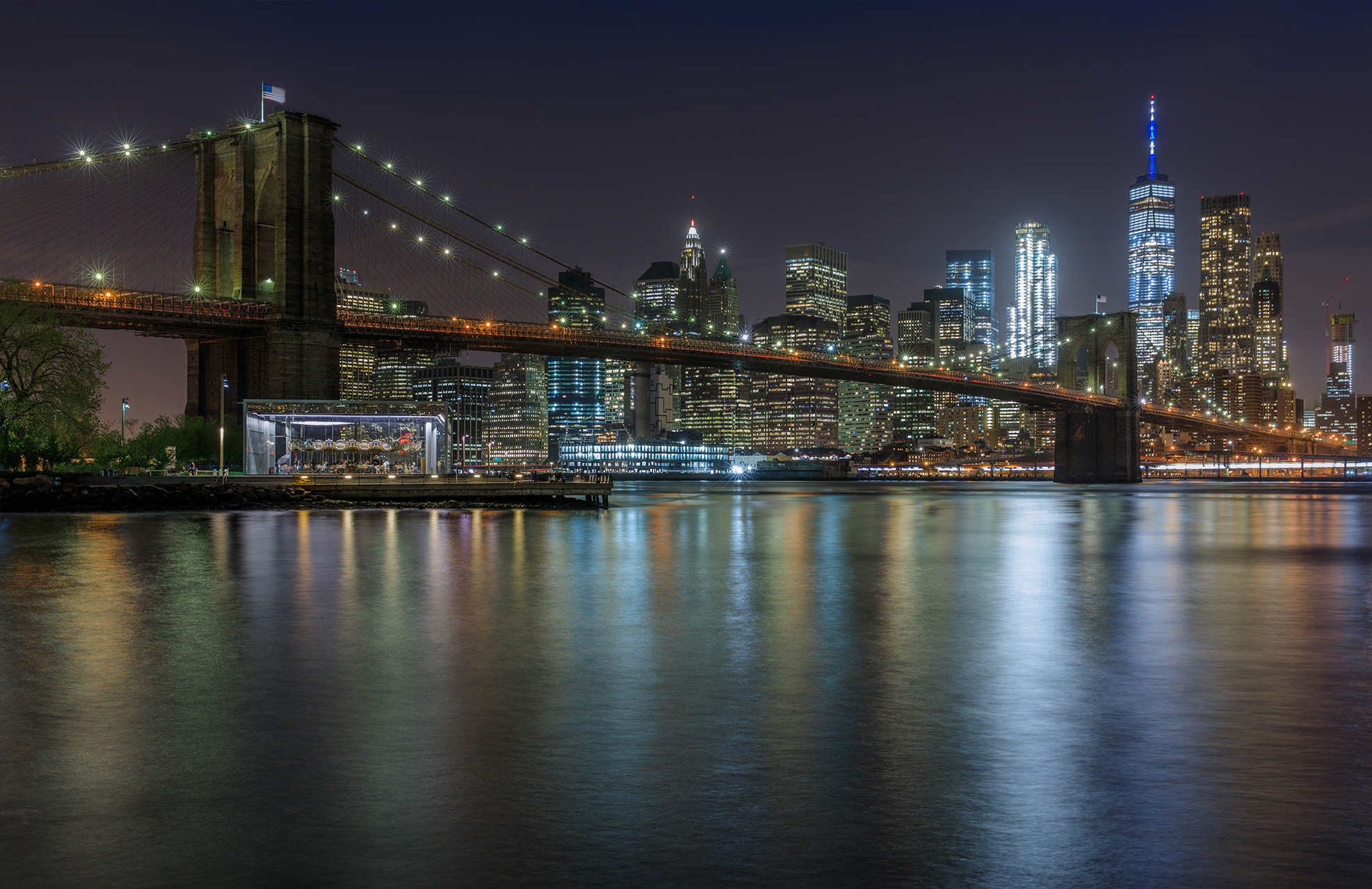 City skyline at night with buildings lit up and a bridge in the foreground.