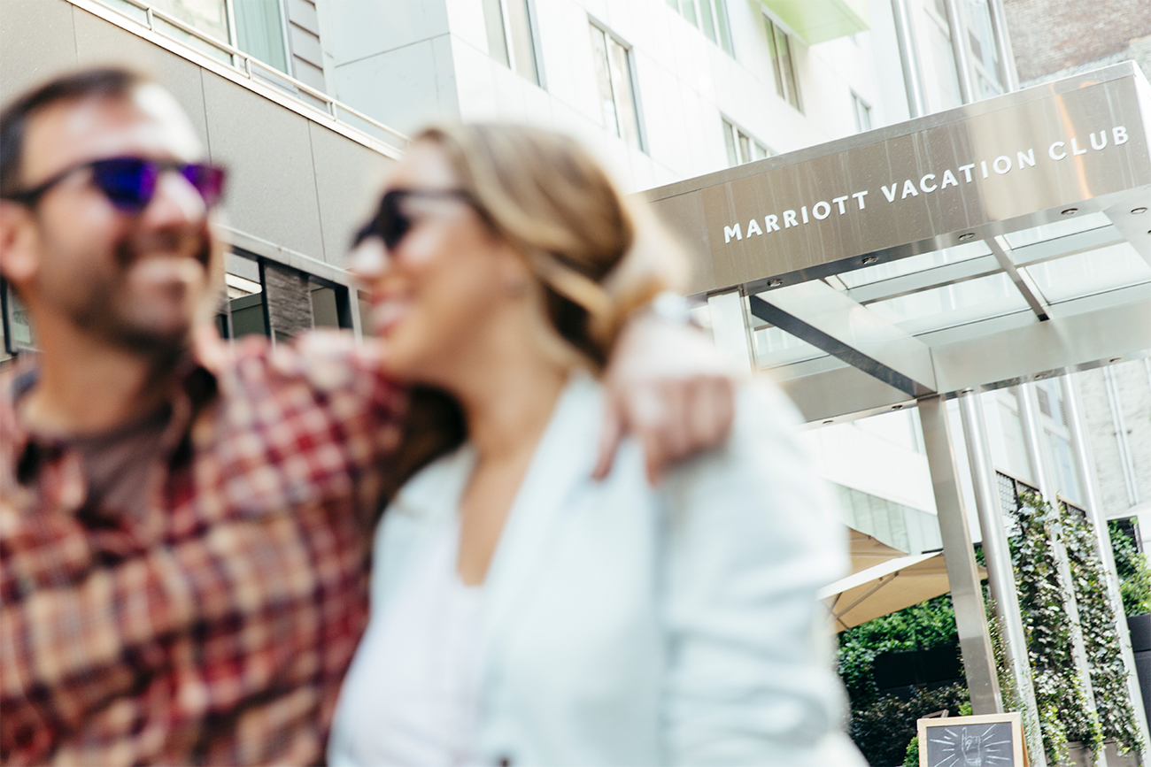 A smiling couple wearing sunglasses step out from the hotel ready to start their day