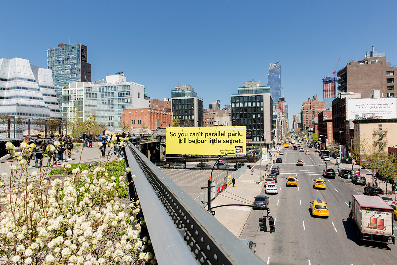 People walk on an elevated walkway over a busy street with the New York skyline in the background