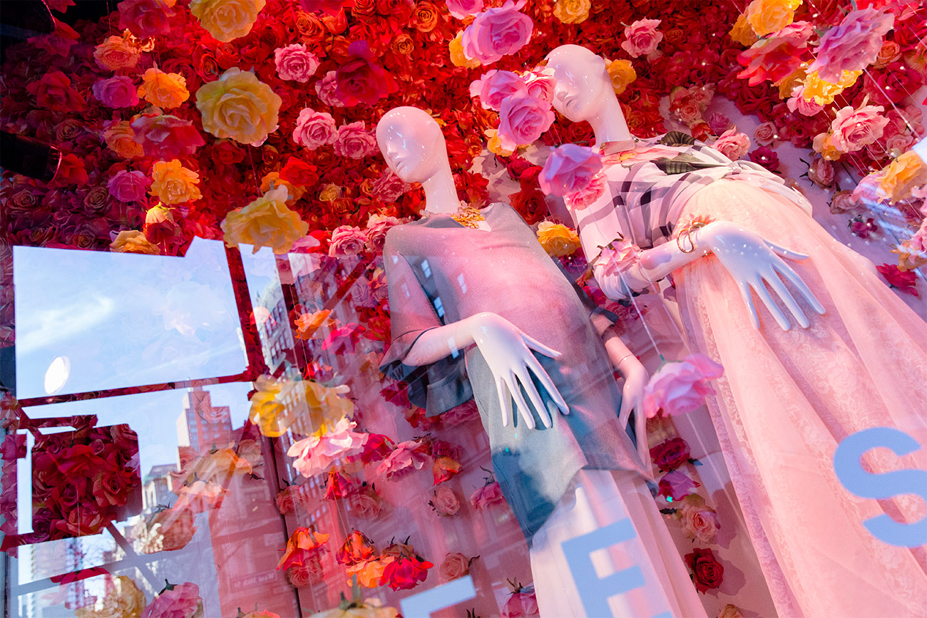 Two mannequins pose in a window display surrounded by colorful flowers