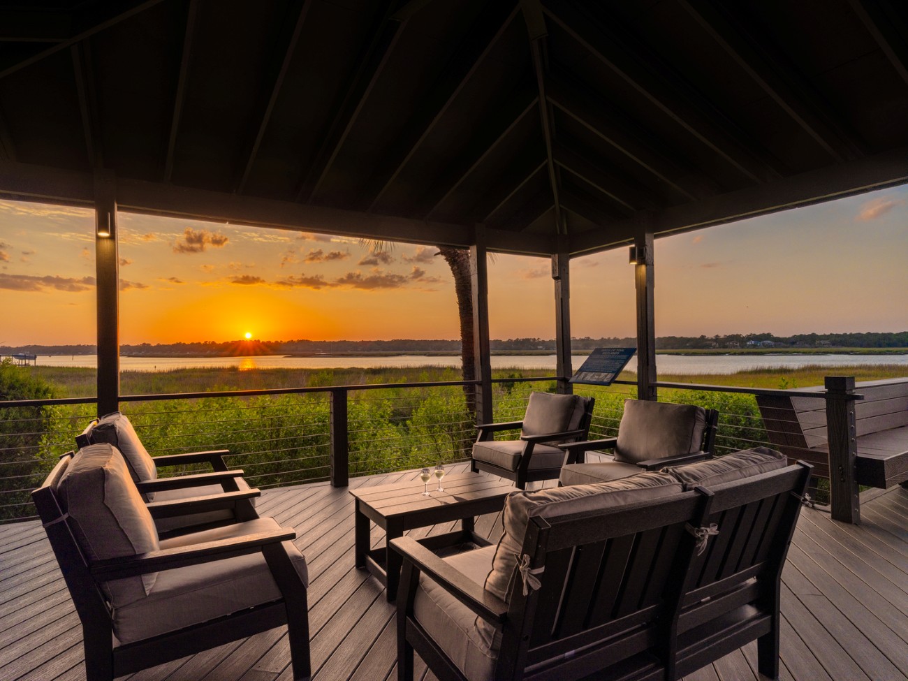 3. Two glasses of wine sit on a table in between seating on a deck overlooking the marsh as the sun begins to set.