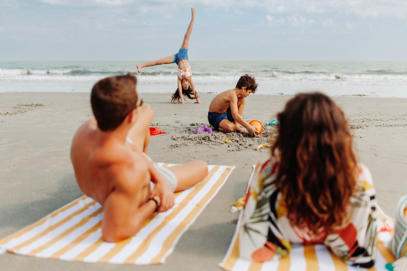 6. A young girl does a cartwheel on the beach as a boy plays in the sand and a couple sit on towels overlooking the ocean. 