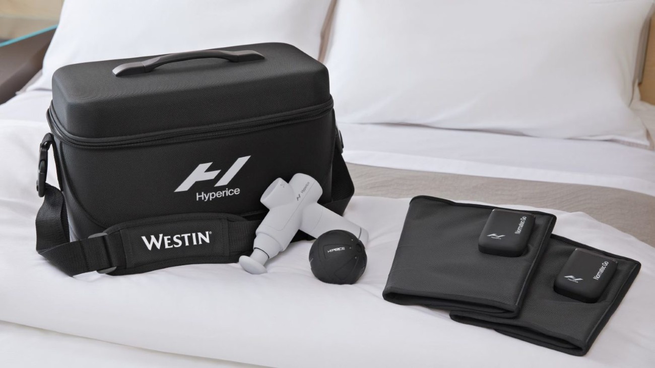 Black travel bag with recovery fitness tools on white bed.