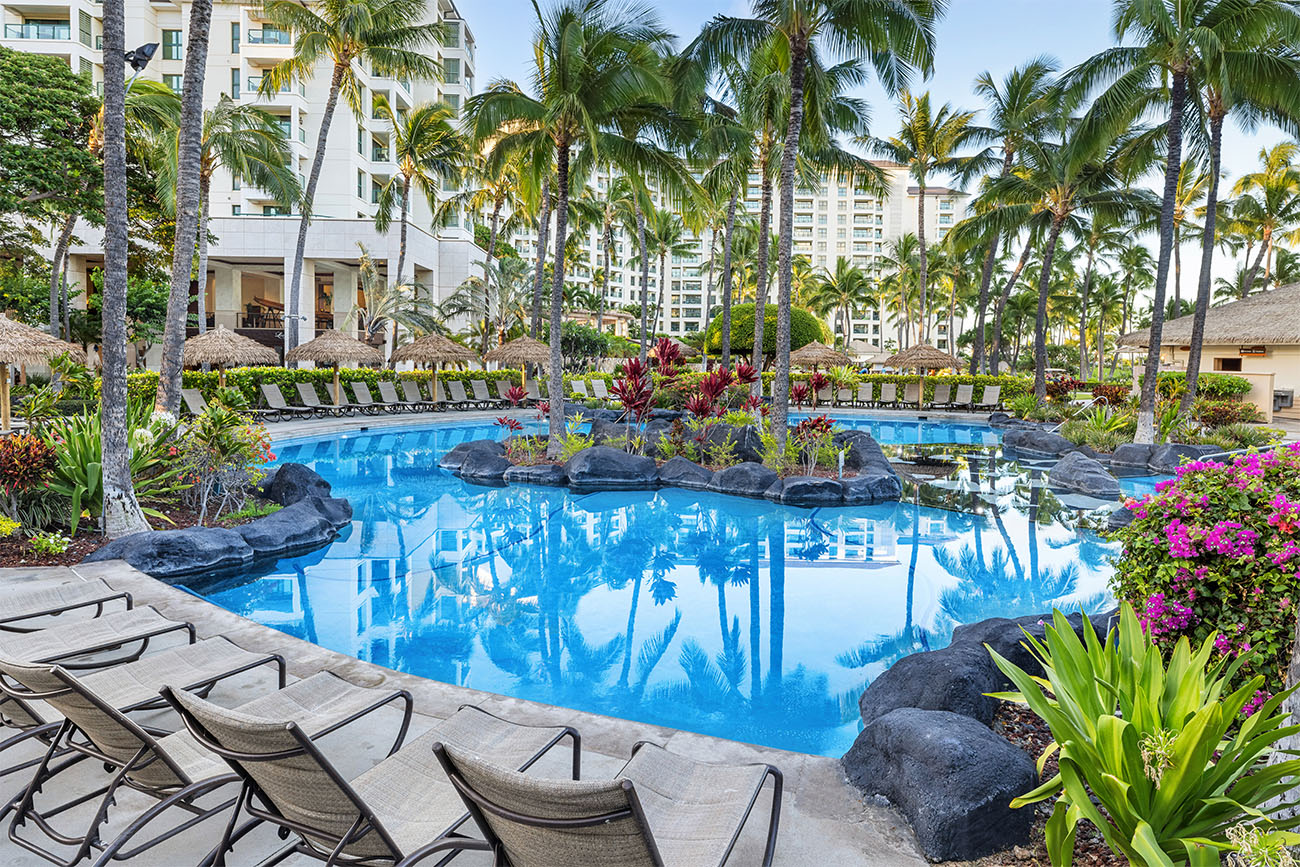 Palm trees and deck chairs are reflected in a calm swimming pool under clear skies.