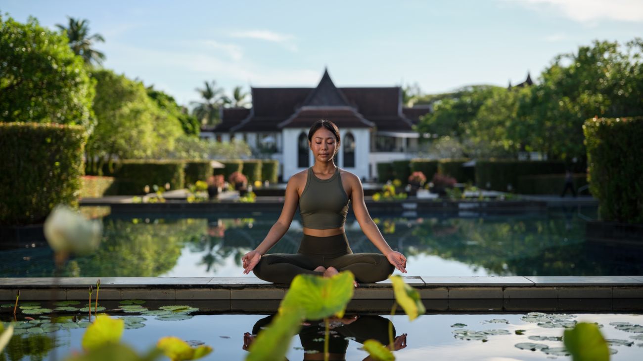 A woman sits in lotus position in front of a Thai building on serene pool.