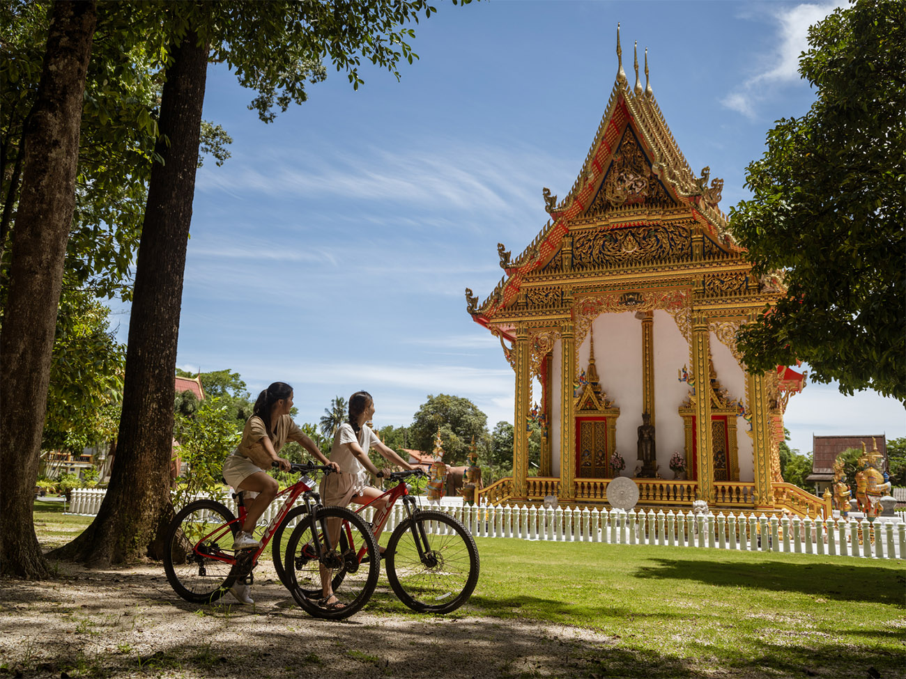 Two people ride bicycles past a Thai temple on a sunny day. 