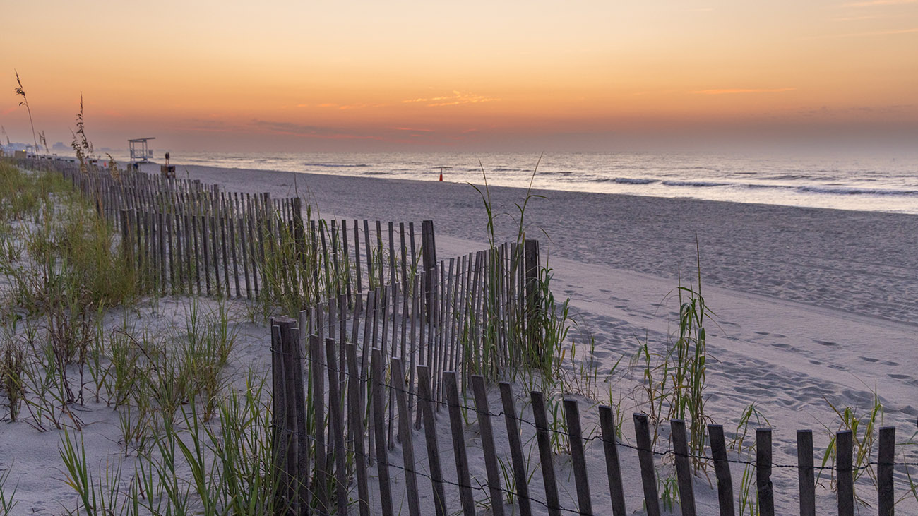 A twilight view of a wooden fence along the dunes of Myrtle Beach, South Carolina, showing a wide sandy shoreline and ocean waves.