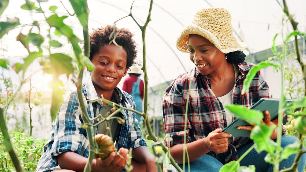 A mother and child tend tomato plants in a greenhouse.