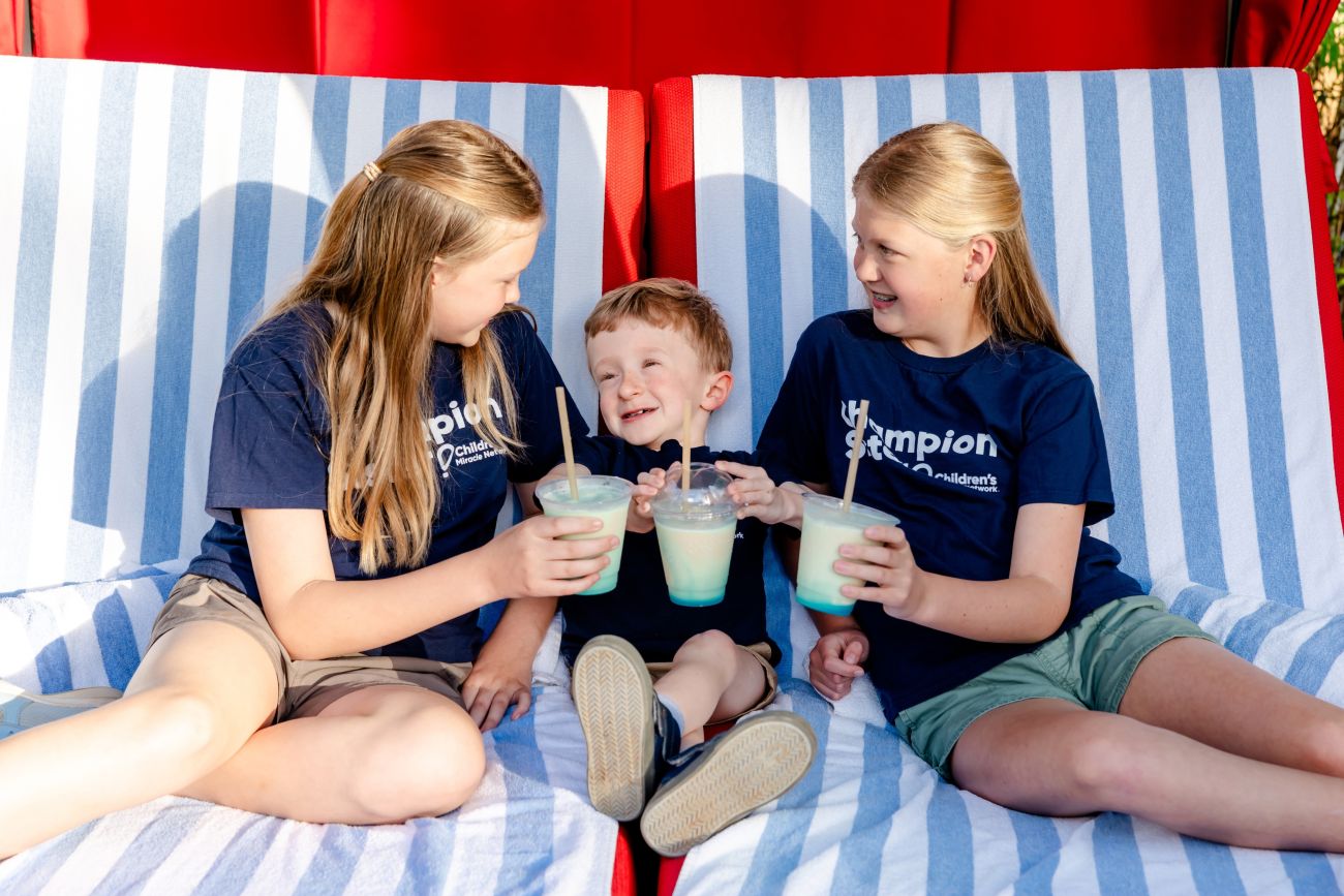 Three children relax on poolside chairs while holding cold smoothies.