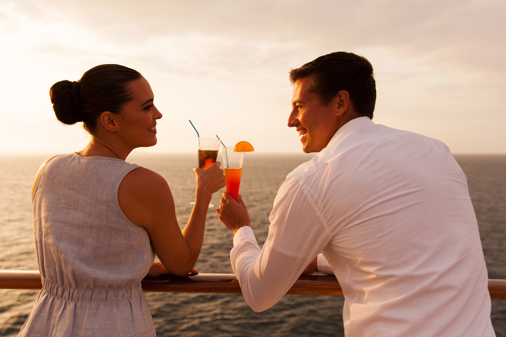 A couple hold drinks on a cruise during sunset.