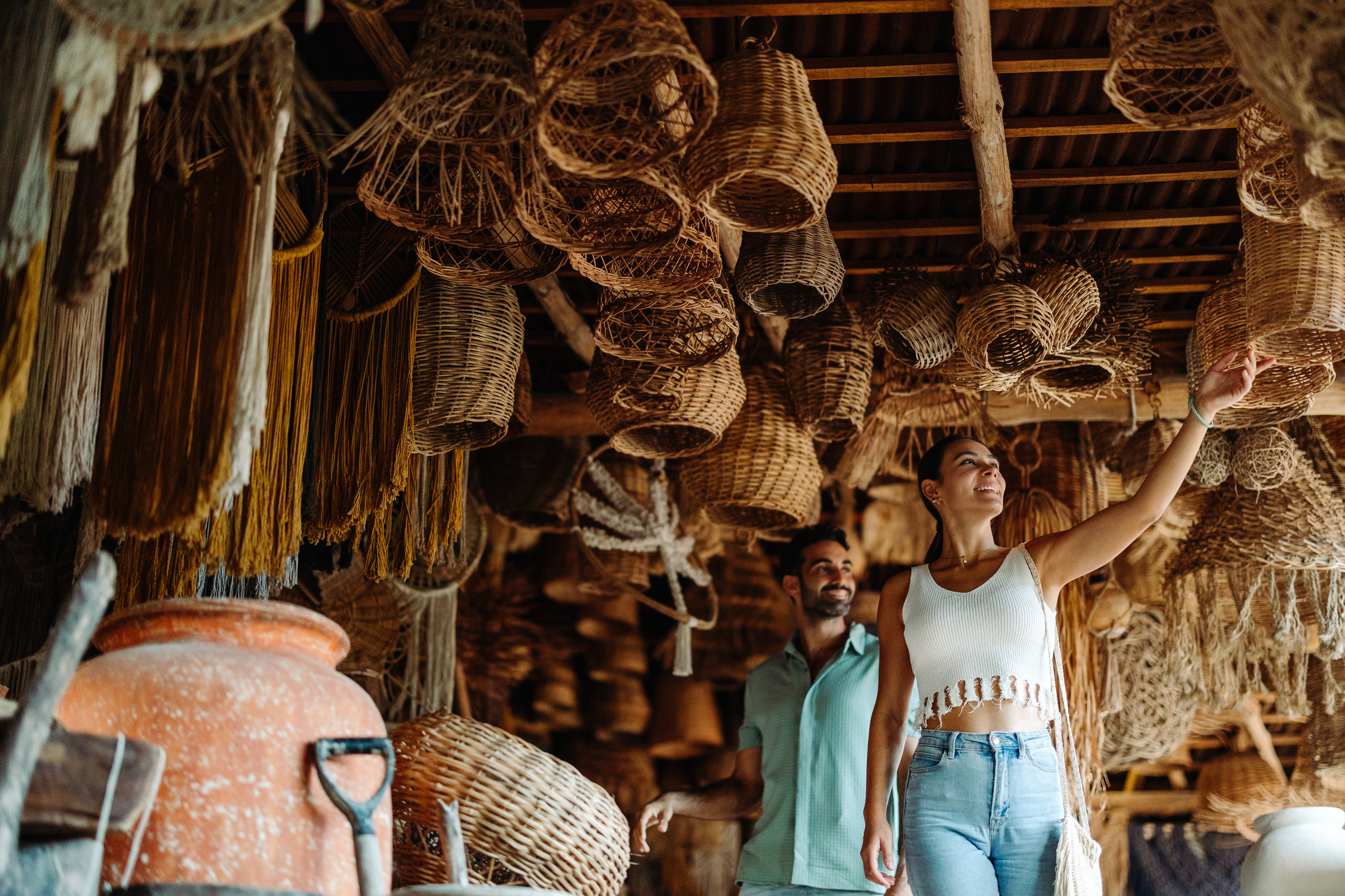Two shoppers browse a shop with baskets hanging from the ceiling and a large clay jar in the foreground.