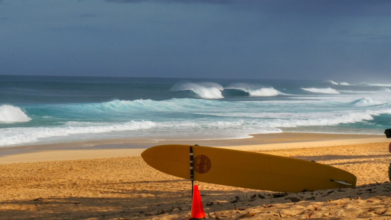A surfboard rests upright on a beach, with waves rolling in along the shoreline.